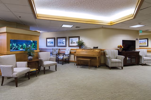 A spacious senior living lounge with armchairs, a piano, a television cabinet, and a built-in fish tank.
