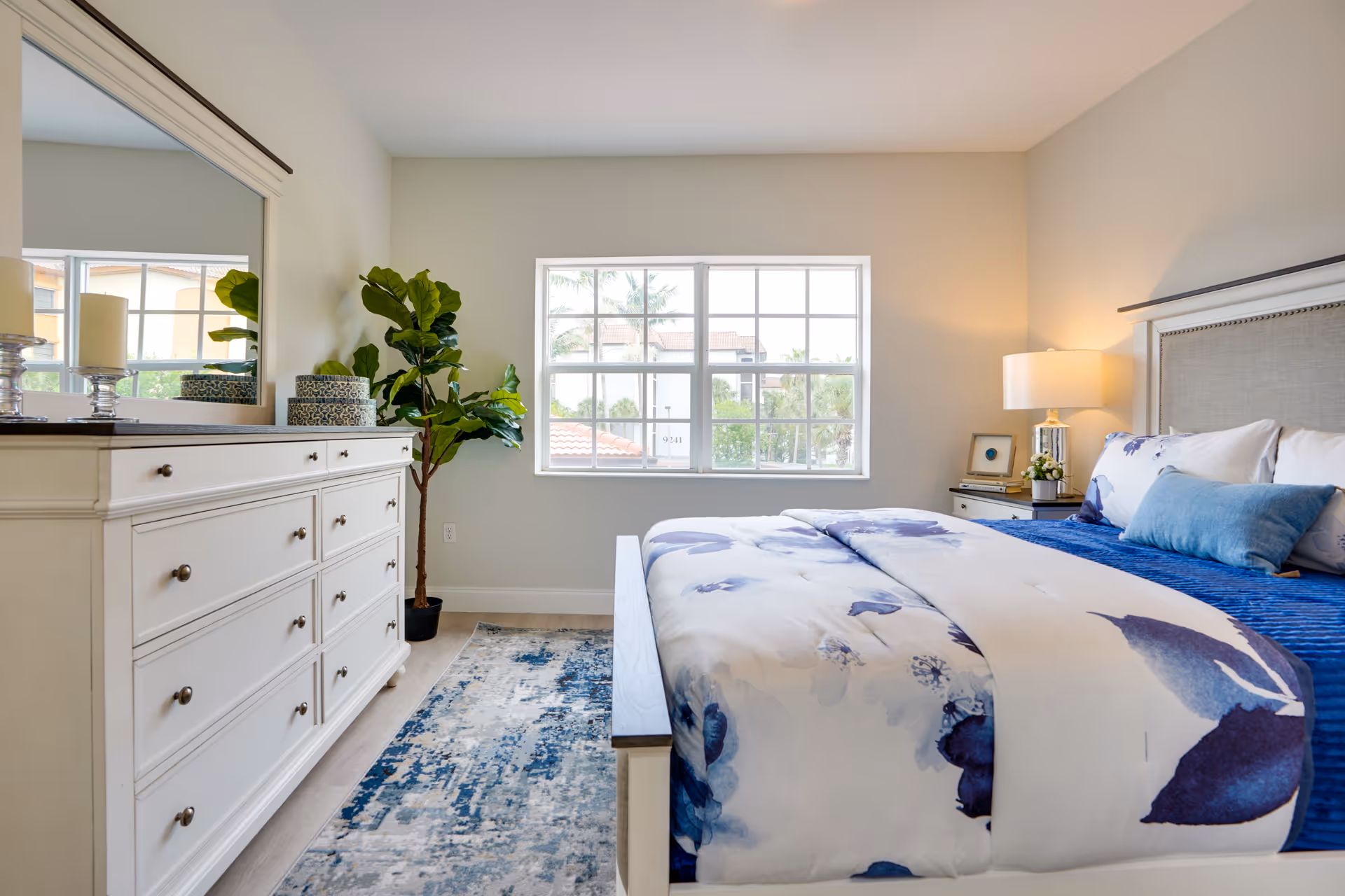 A bright and airy bedroom with a large window letting in natural light. The room features a bed with white and blue floral bedding, a white dresser with a large mirror above it, a bedside table with a lamp, and a green potted plant in the corner. A blue and white rug is placed on the floor beside the bed.
