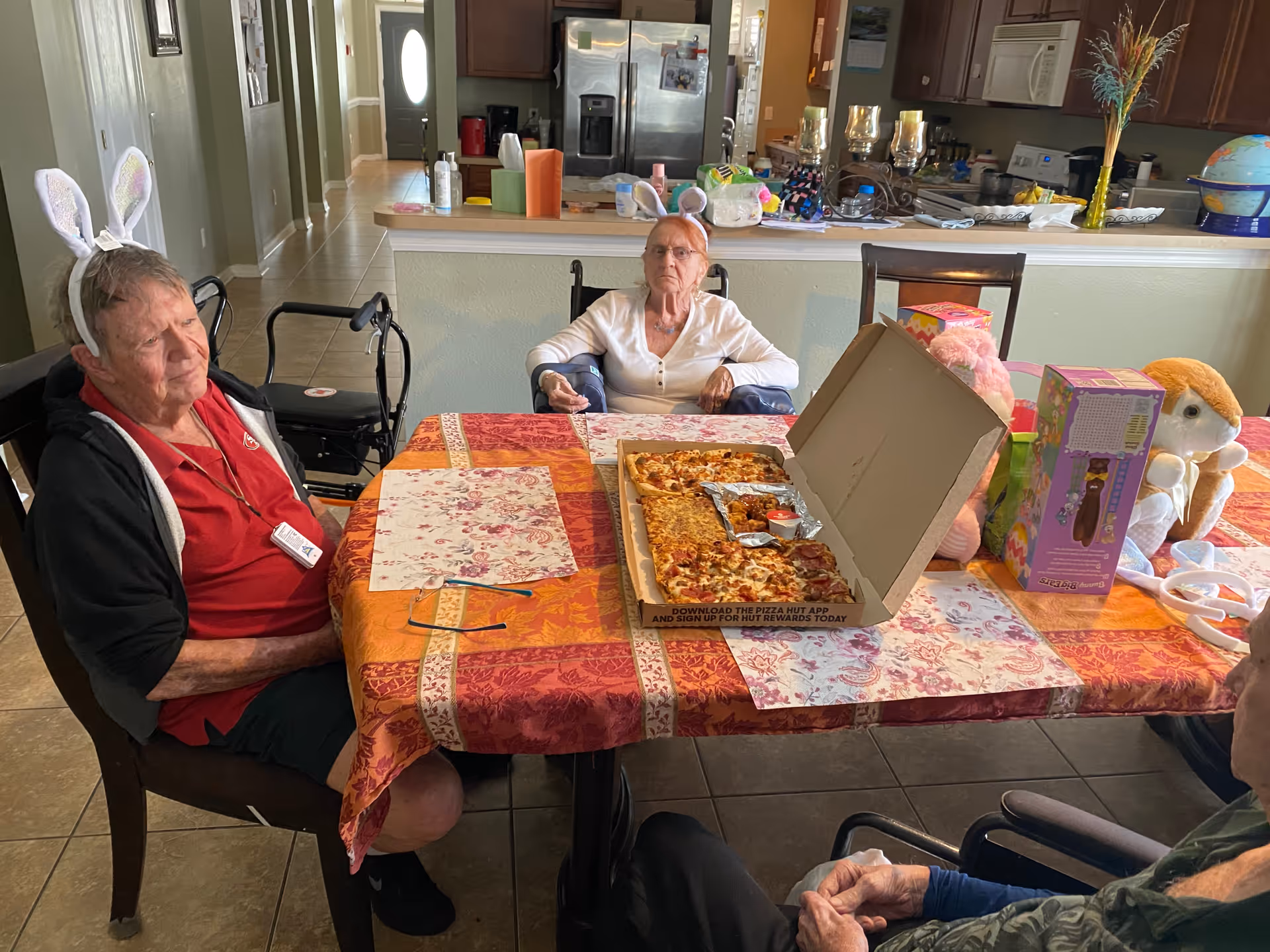 Three elderly individuals sitting around a dining table with a pizza box containing various types of pizza. Two of the individuals are wearing bunny ear headbands. The table is covered with a colorful tablecloth and has some toys and boxes on it. The background shows a kitchen area with cabinets, a refrigerator, and various kitchen items.