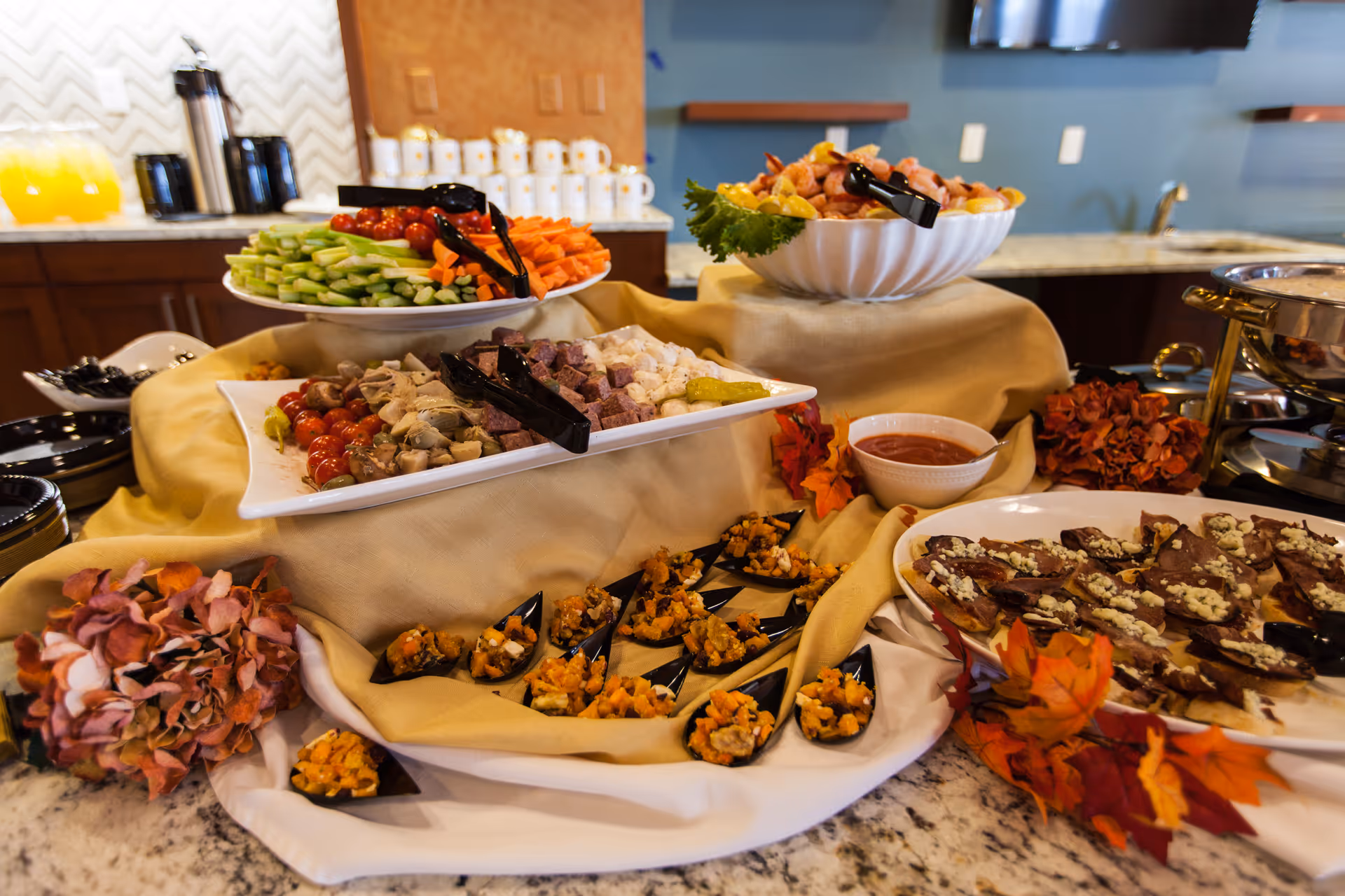 A buffet table with a variety of appetizers including sliced vegetables, cubed meats and cheeses, small servings of mixed dishes in black spoons, and a plate of crackers topped with cheese. The table is decorated with autumn leaves and flowers, and there are coffee cups and a beverage dispenser in the background.