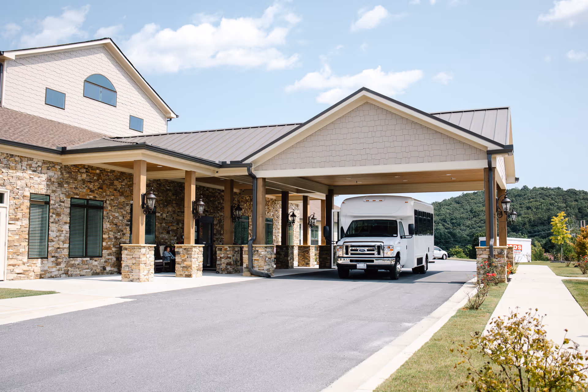 Exterior view of Dahlonega Assisted Living & Memory Care facility showing a covered entrance with stone pillars and a white shuttle bus parked underneath. The building has a stone facade with several windows and a clear sky with some clouds in the background.