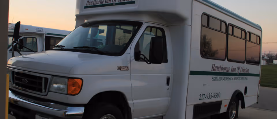 White shuttle bus with the text 'Hawthorne Inn Of Clinton Skilled Nursing & Assisted Living' and a phone number parked outdoors during sunset.