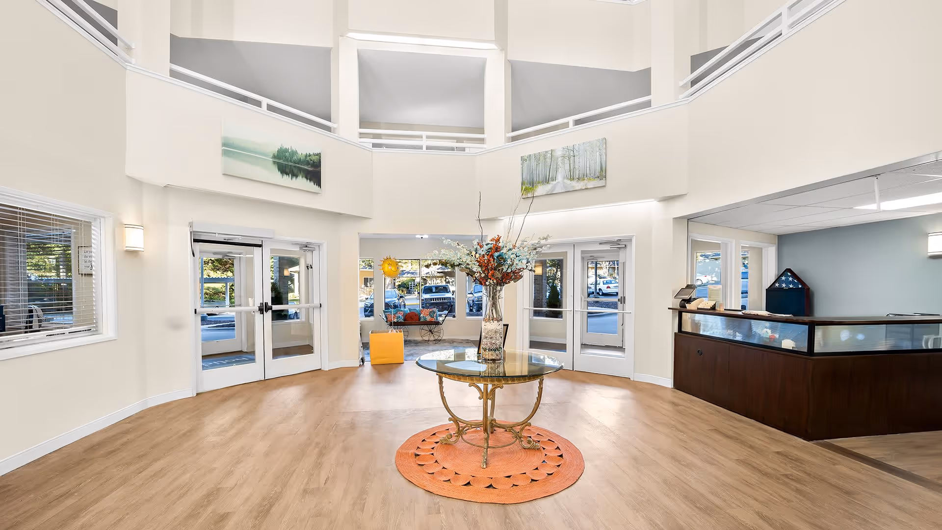 Spacious and bright lobby area of Holiday Durham Regent with light wood flooring, a round glass table with a floral arrangement on an orange rug in the center, double glass doors leading outside, a reception desk to the right, and large windows allowing natural light.