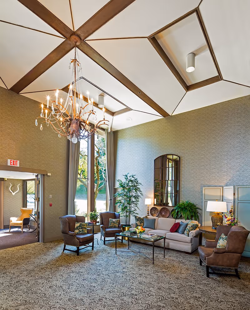 Bright, elegant communal living room with a high coffered ceiling and chandelier, tall windows, sofas and armchairs arranged around a coffee table.