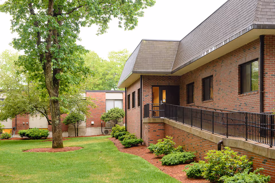 Brick exterior of Life Care Center of Acton showing a ramped entrance, windows, and landscaped lawn with trees and shrubs.