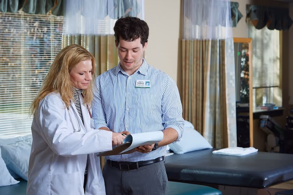 A female doctor in a white coat and a male staff member with a name tag are standing together in a medical or therapy room, looking at and discussing a clipboard. The room has treatment tables, large windows with blinds, and curtains.
