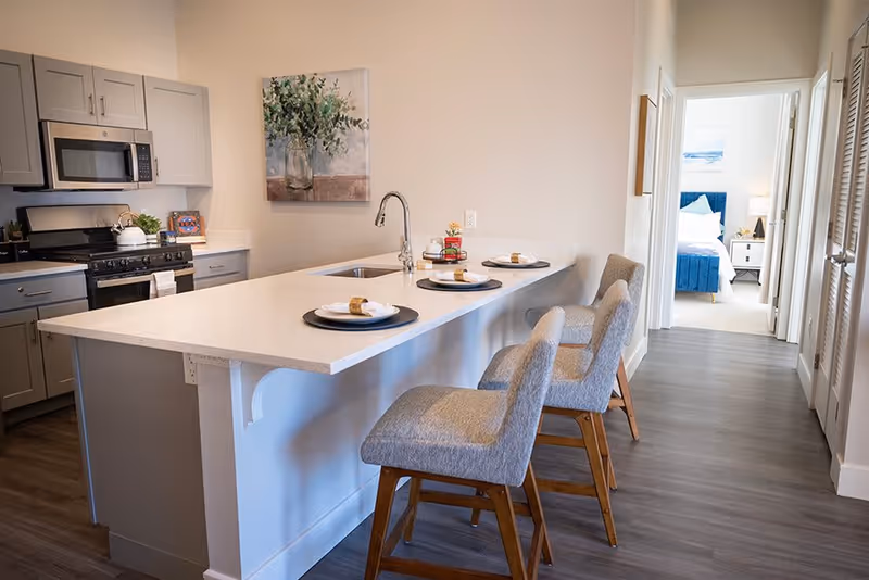 Bright modern kitchen with a white island countertop, three upholstered bar stools, and a view into a bedroom.