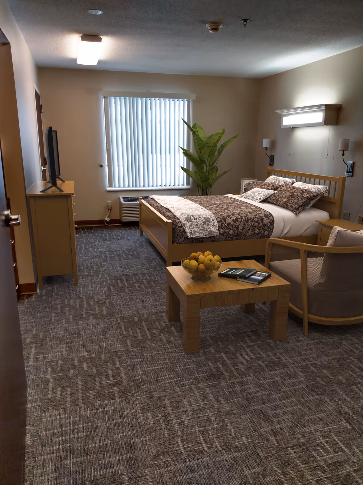 A cozy bedroom in an assisted living facility featuring a bed with patterned bedding, a wooden nightstand, a small wooden table with a bowl of fruit and brochures, a cushioned chair, a TV on a dresser, a large window with vertical blinds, and a tall green plant in the corner.