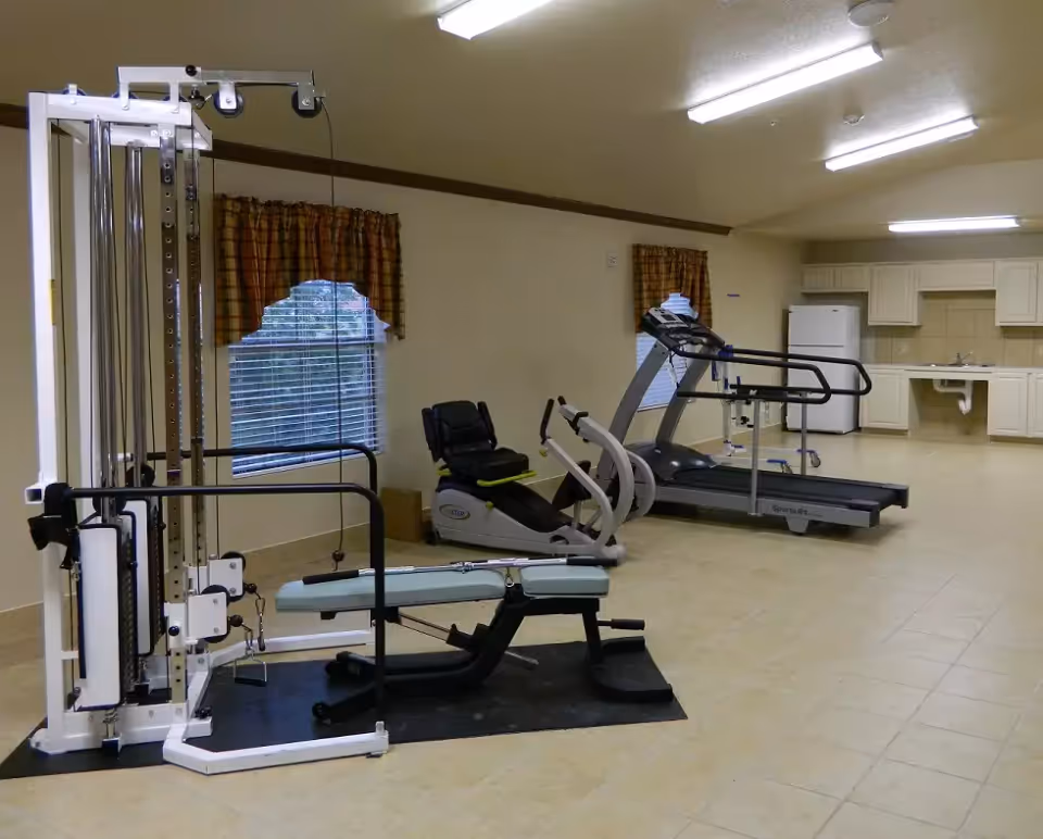 A fitness room with a weight machine and bench in the foreground, a recumbent bike and treadmill in the middle, and a small kitchenette along the back wall.