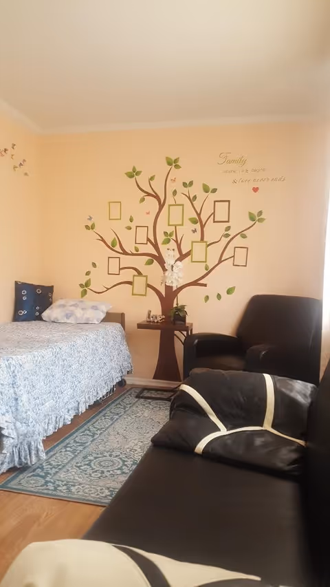 A cozy room with a single bed covered in a blue patterned bedspread and a pillow. Next to the bed is a small table with a white orchid plant and a decorative 'love' sign. On the wall behind the bed is a large tree decal with empty picture frames and green leaves, along with the quote 'Family where life begins & love never ends.' There is a black armchair and a black couch with patterned cushions in the foreground. The floor has a blue and white patterned rug.