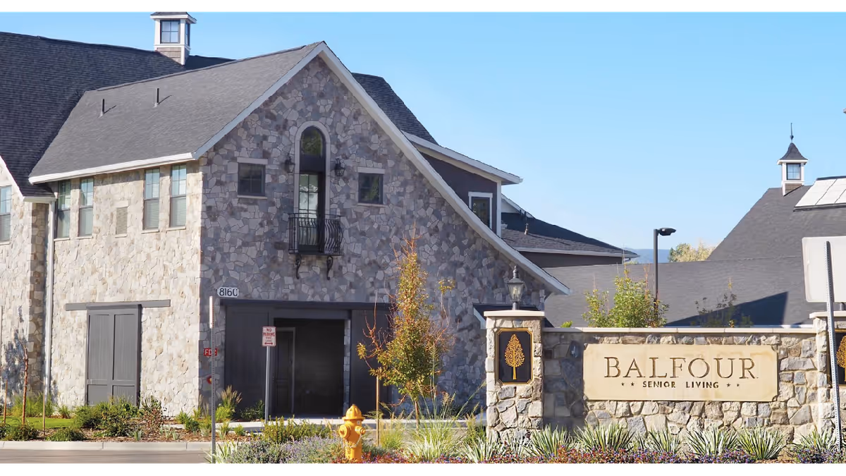 Exterior view of Balfour Littleton senior living facility showing a stone building with a pitched roof, windows, and a small balcony. In front, there is a stone sign with the text 'BALFOUR SENIOR LIVING' and decorative landscaping including plants and a yellow fire hydrant.