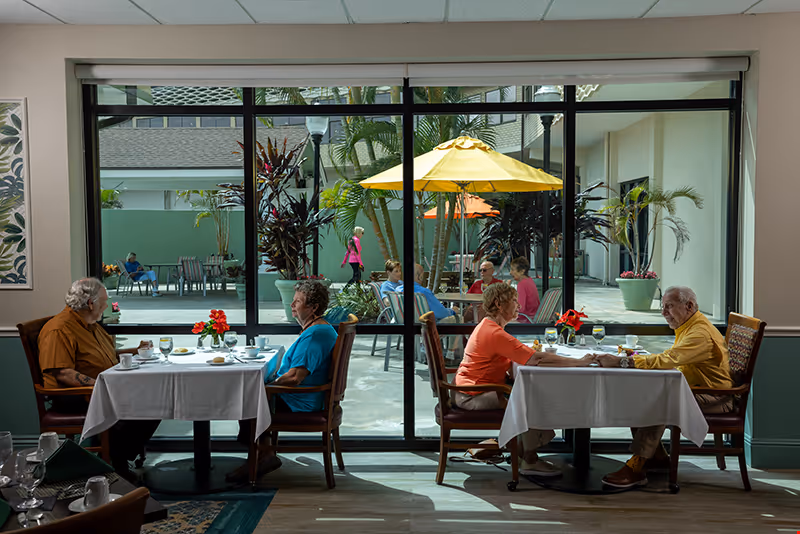 Two pairs of senior residents sit at dining tables inside a dining room with a large window showing a courtyard with umbrellas and other people.