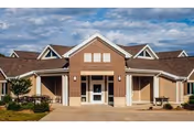 Front entrance of a single-story brick and siding senior living building with a covered portico, peaked rooflines, walkway, and small landscaped areas under a blue sky.