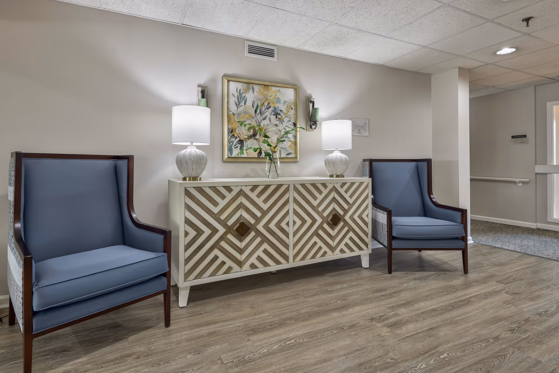 Lobby seating area with two blue armchairs flanking a patterned console table topped with lamps and floral artwork.