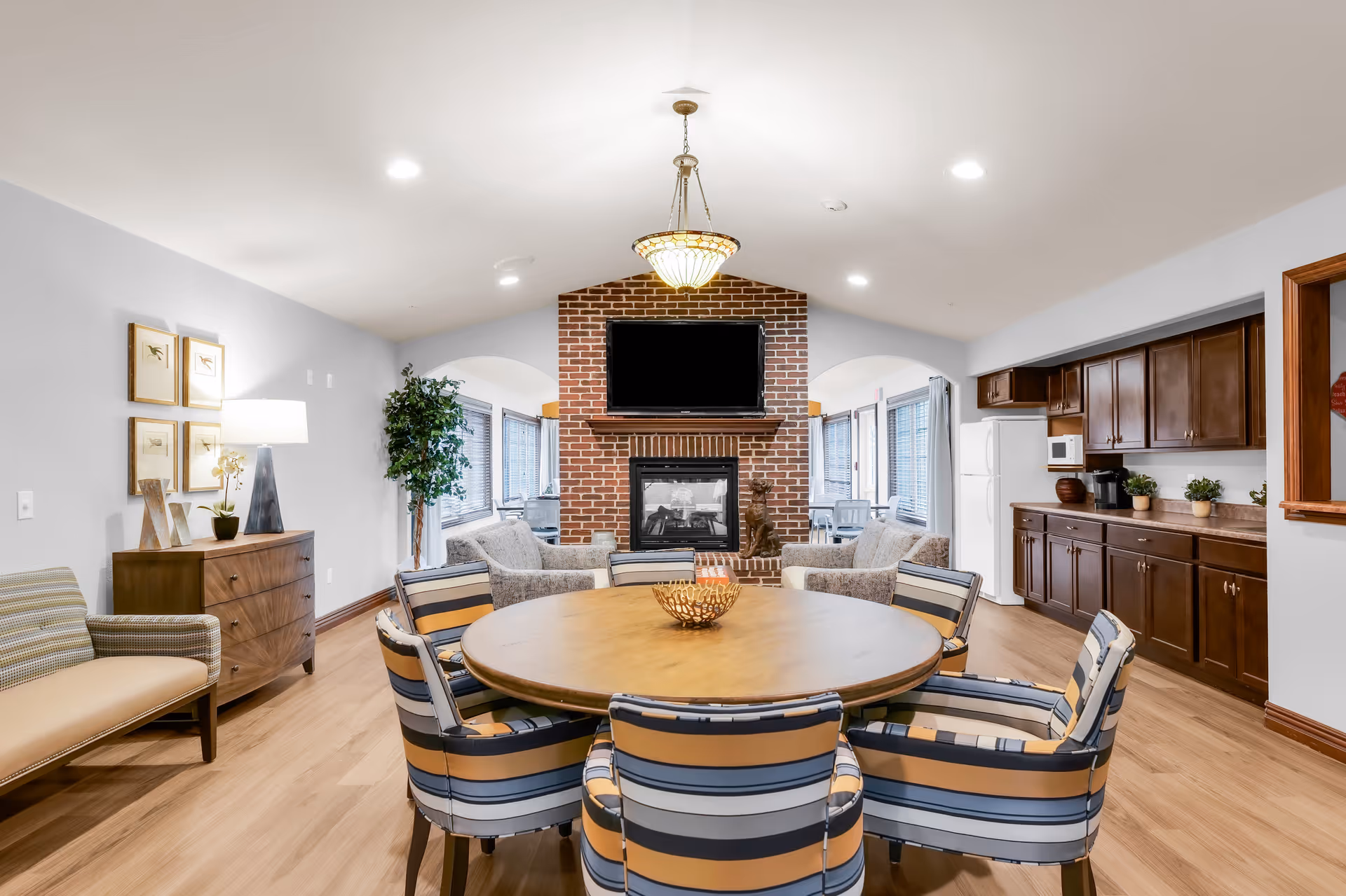 Bright communal living and dining area with a round table and striped chairs facing a brick fireplace and wall-mounted TV, with a kitchenette along the right wall.