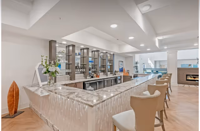 Bright modern indoor bar and lounge area with a marble countertop, bar stools, back shelving with bottles, and seating near a fireplace.