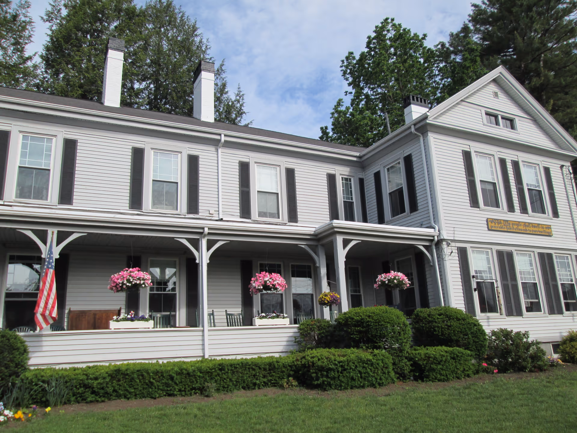 Exterior view of a large two-story white building with black shutters and two chimneys. The building has a covered porch with hanging flower baskets and an American flag. There are well-maintained bushes and a green lawn in front of the building, with tall trees in the background under a partly cloudy sky.