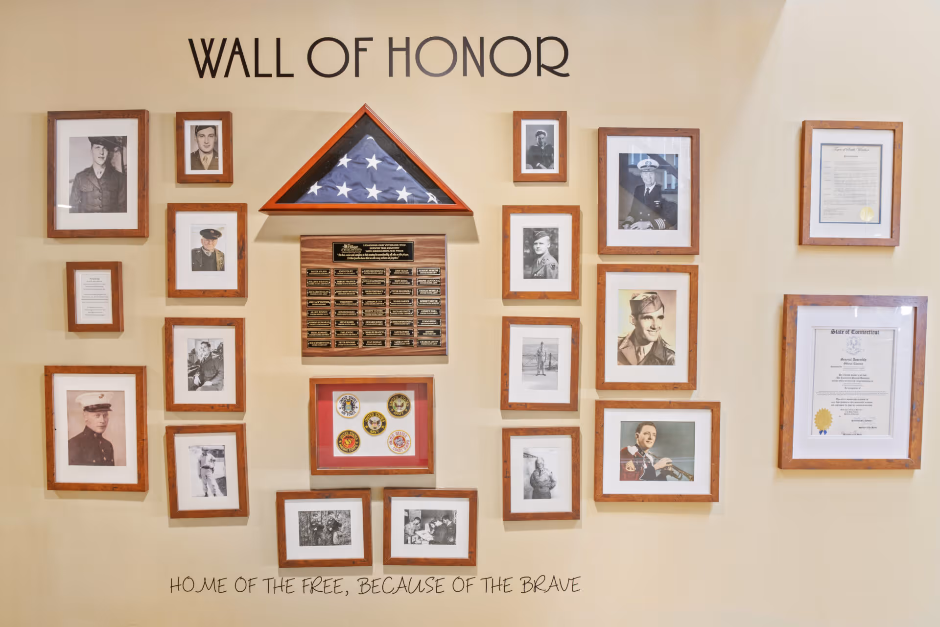 A Wall of Honor display featuring framed photographs of military personnel, a folded American flag in a triangular case, plaques with names, military insignia, and certificates. The wall has the text 'WALL OF HONOR' at the top and 'HOME OF THE FREE, BECAUSE OF THE BRAVE' at the bottom.