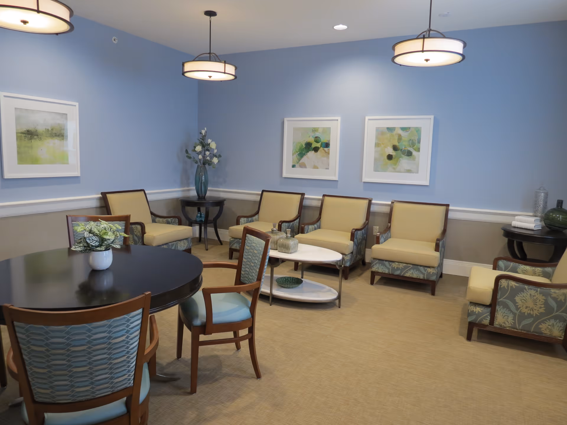 A cozy sitting area in a senior living facility with light blue walls and beige carpet. The room features a round dark wood table with four chairs, a small white coffee table, and six cushioned armchairs with floral upholstery. Three abstract paintings hang on the wall, and two pendant lights illuminate the space. There are decorative vases with flowers on side tables.