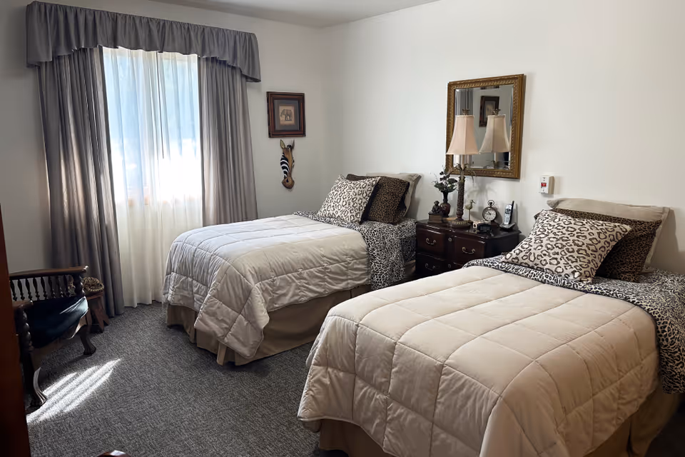 Sunlit twin bedroom with two neatly made beds, a shared nightstand with lamps and mirror, and a curtained window.