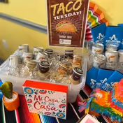 A colorful display featuring a plastic container filled with various drink cans and bottles on ice, a sign that says 'Taco Tuesday!' with a taco illustration, a small decorative sign reading 'Mi Casa es su Casa', a small potted cactus, blue boxes labeled 'Ard Law Firm', and a colorful piñata on a vibrant striped tablecloth.