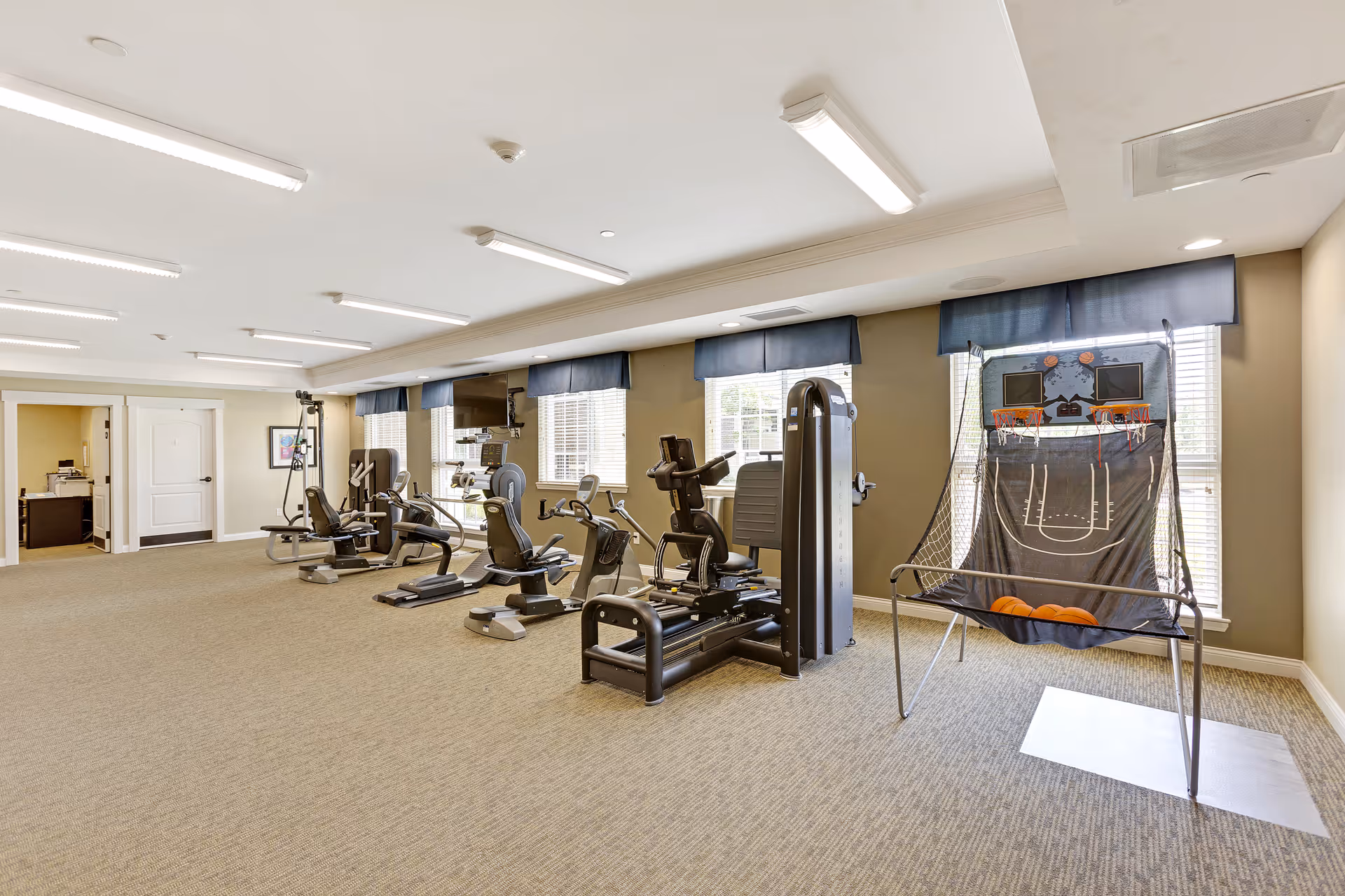 A spacious fitness room with exercise equipment including stationary bikes, weight machines, and a basketball shooting game near windows with blue valances. The room has beige walls, carpeted floor, and bright overhead lighting.