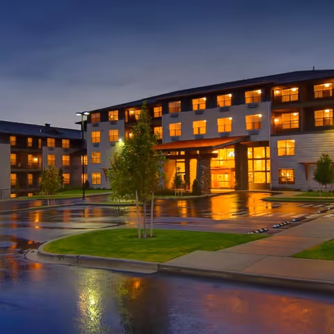 Exterior view of a multi-story senior living facility at dusk with lights glowing warmly from the windows. The building is surrounded by a wet parking lot and landscaped areas with small trees and grass.