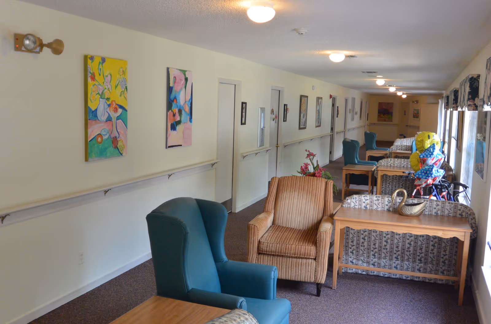 A long, narrow common area in a senior living facility with several armchairs and small tables arranged along the wall and windows. The walls are decorated with colorful paintings, and there are handrails along the wall. The room is well-lit with ceiling lights and natural light from the windows on the right side.