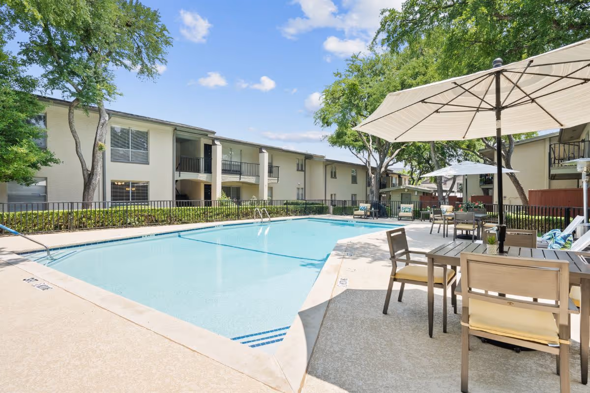 Outdoor swimming pool area at Meadowstone Place with clear blue water, surrounded by a concrete deck. There are tables and chairs with large umbrellas providing shade. The pool area is fenced and there are two-story residential buildings and trees in the background under a blue sky with some clouds.