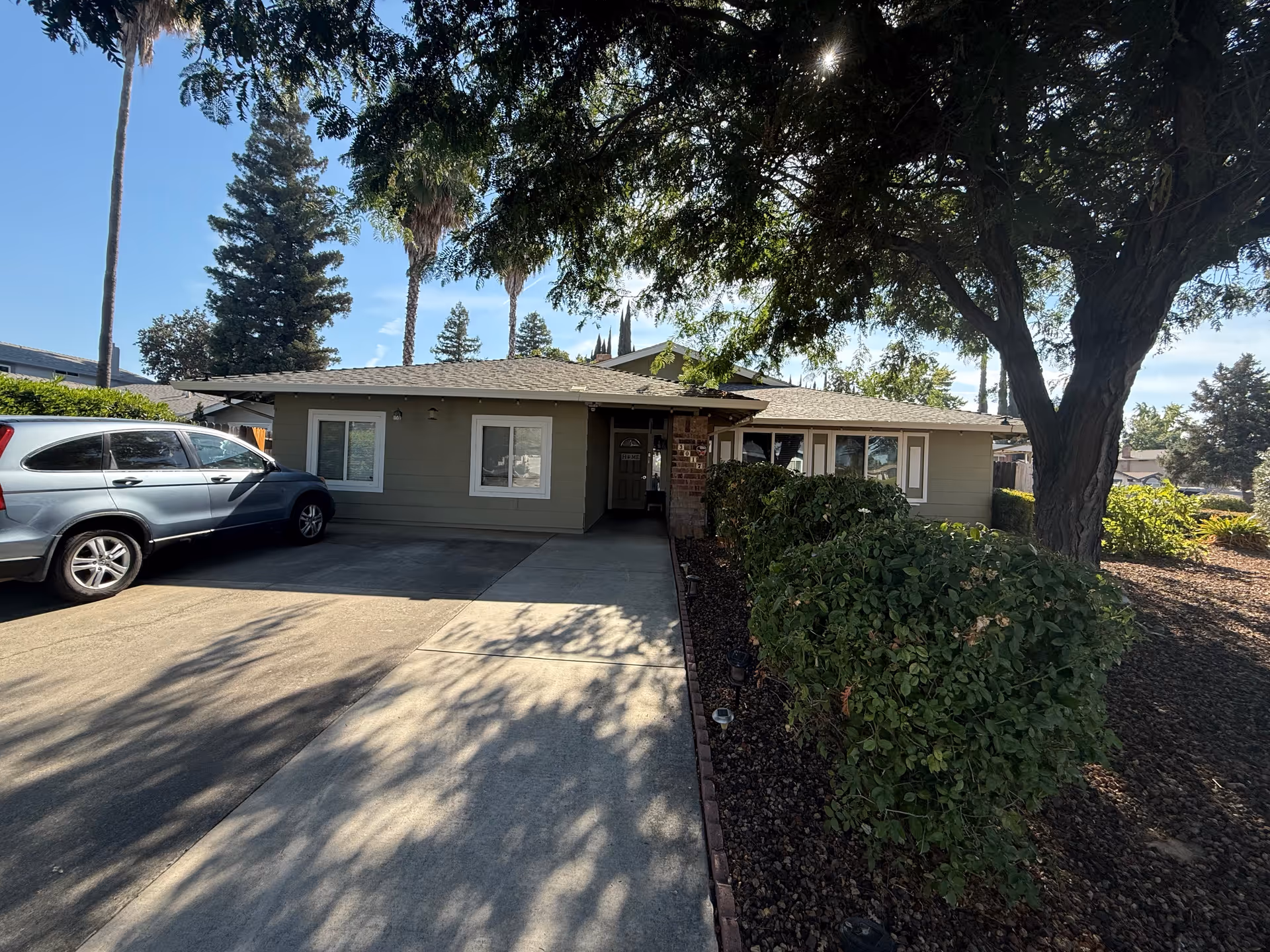 Single-story house front with a driveway, a parked SUV, hedges, and a large tree casting shade.
