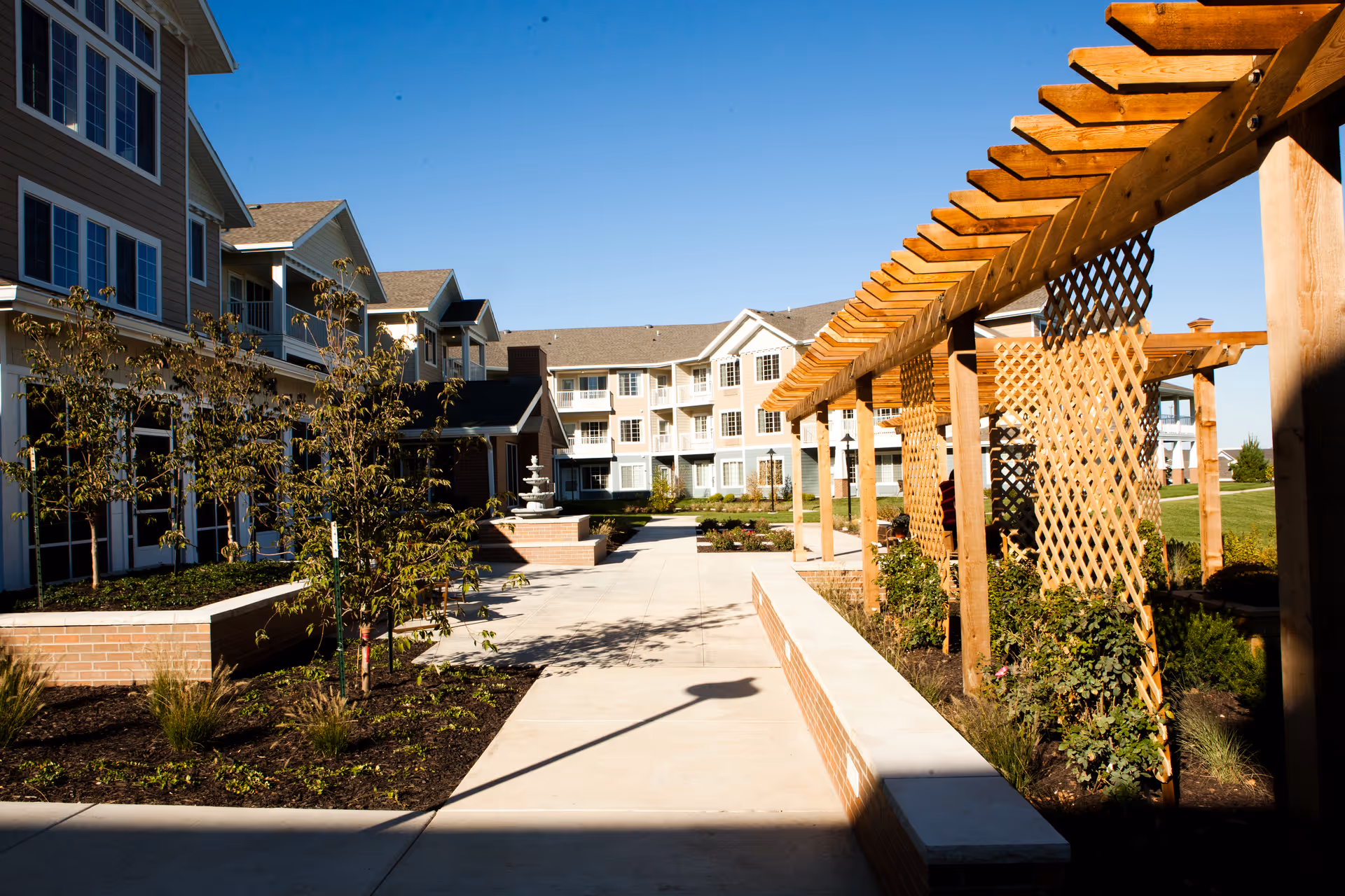 Outdoor courtyard area of Meadowlark Estates Gracious Retirement Living featuring a paved walkway, landscaped garden beds with small trees and shrubs, a wooden pergola on the right side, and multi-story residential buildings in the background under a clear blue sky.