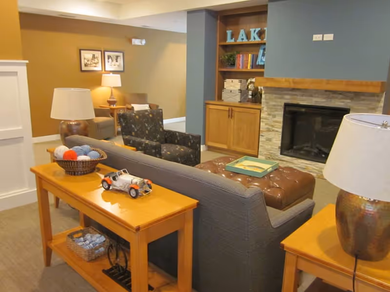 Community lounge with sofas and armchairs facing a fireplace, built-in shelving, ottoman and side tables with lamps.