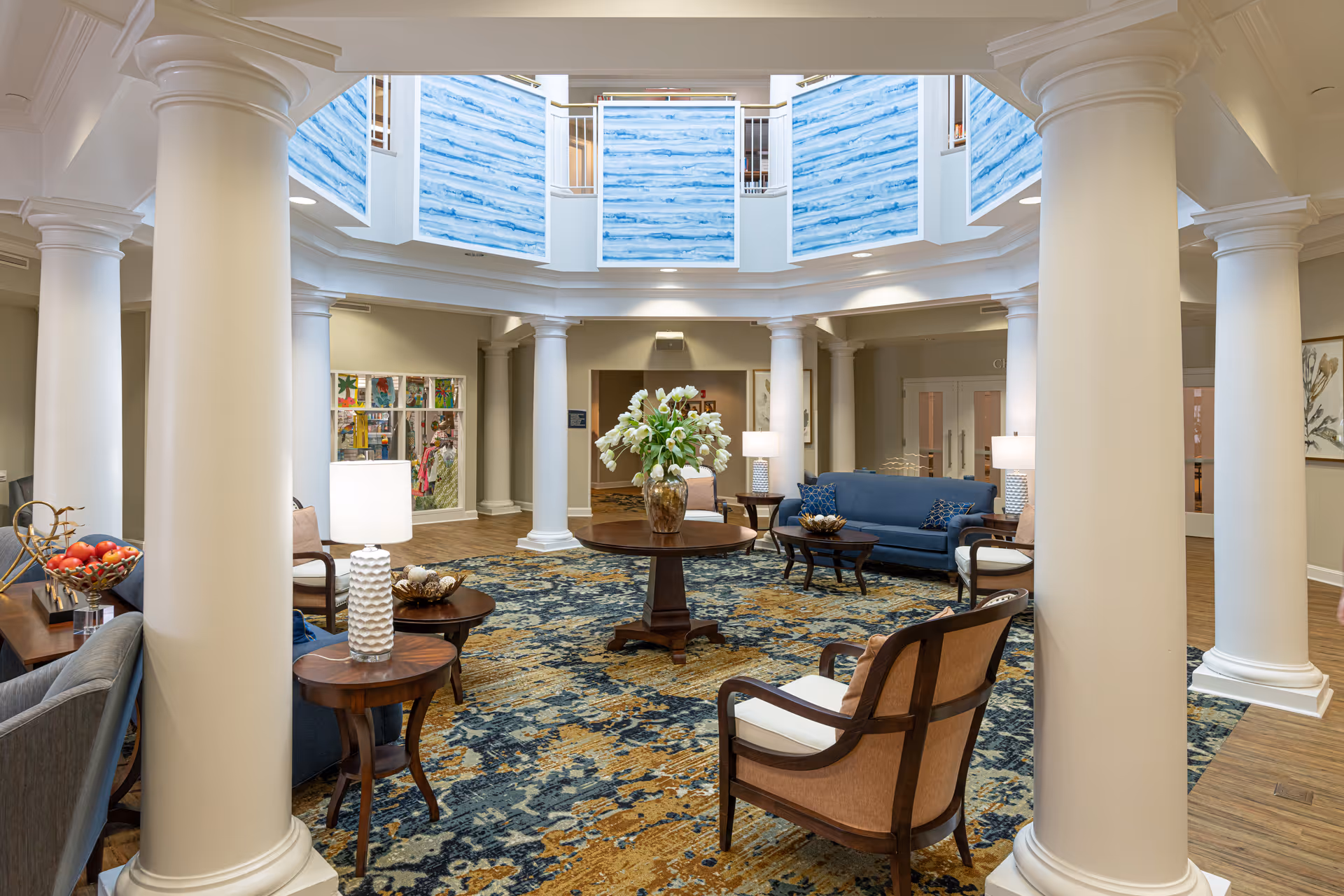 A spacious and elegant living room area with large white columns, a patterned blue and gold carpet, several wooden chairs with cushions, a blue sofa, side tables with lamps, and a round wooden table with a vase of white flowers in the center. The room has a high ceiling with blue and white artwork panels and warm lighting.