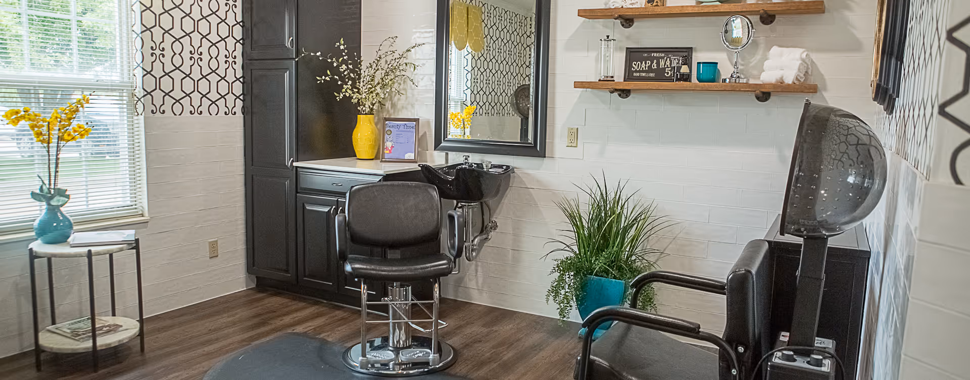 A bright and clean hair salon area with a black salon chair in front of a sink and mirror. The room features dark wood cabinets, decorative shelves with towels and small items, a potted plant, and a hair dryer chair. There is a window with blinds and a small table holding a vase with yellow flowers.