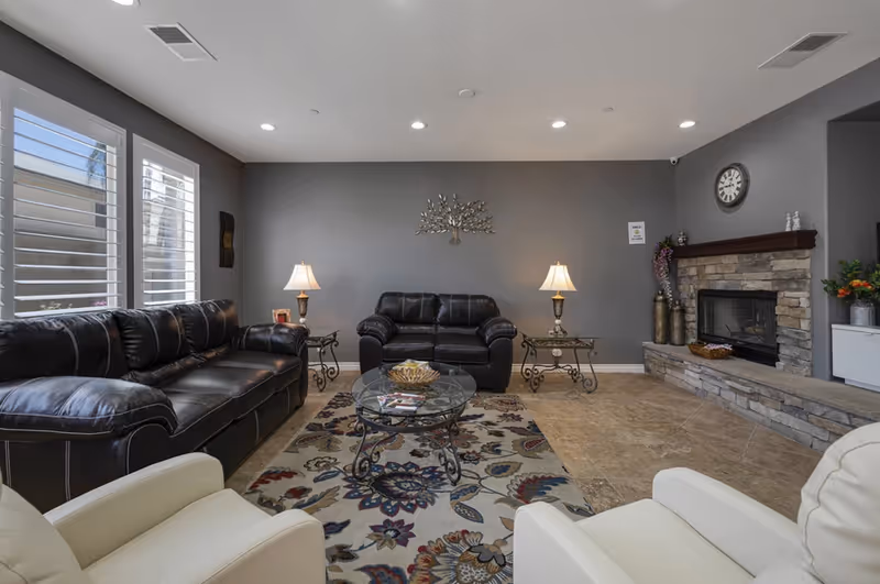 A cozy senior living room with two black leather sofas, two white armchairs, a glass coffee table on a floral patterned rug, two side tables with lamps, a stone fireplace with a clock above it, and a decorative metal tree wall art on a gray wall.
