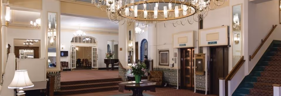 Elegant retirement residence lobby with chandeliers, seating areas, a central table with flowers, elevator doors and a carpeted staircase.