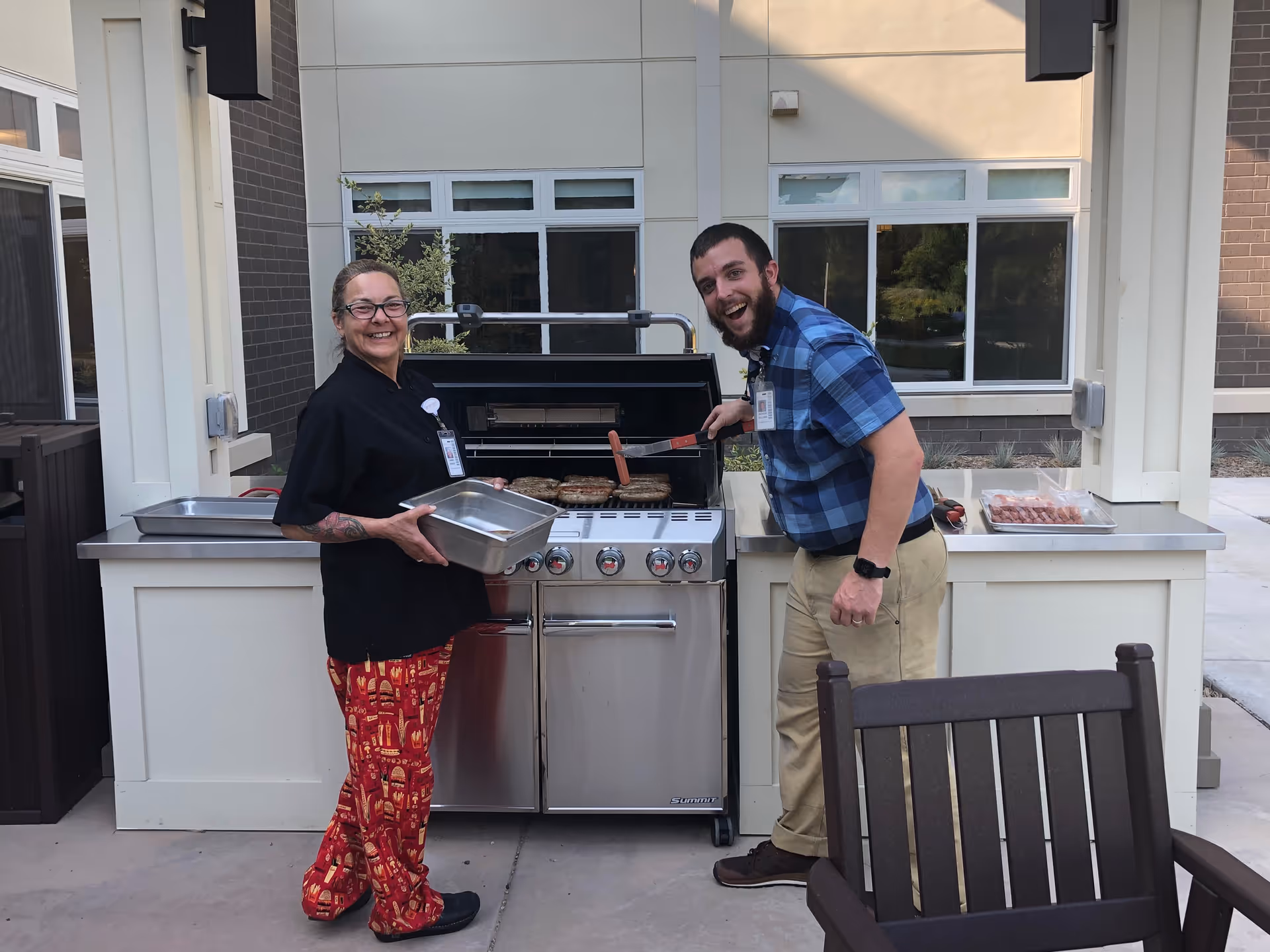 Two staff members smiling and grilling food on an outdoor gas grill at a senior living facility. One person is holding a metal tray, and the other is using a spatula to cook sausages and patties. They are standing in a covered outdoor area with windows and a chair nearby.