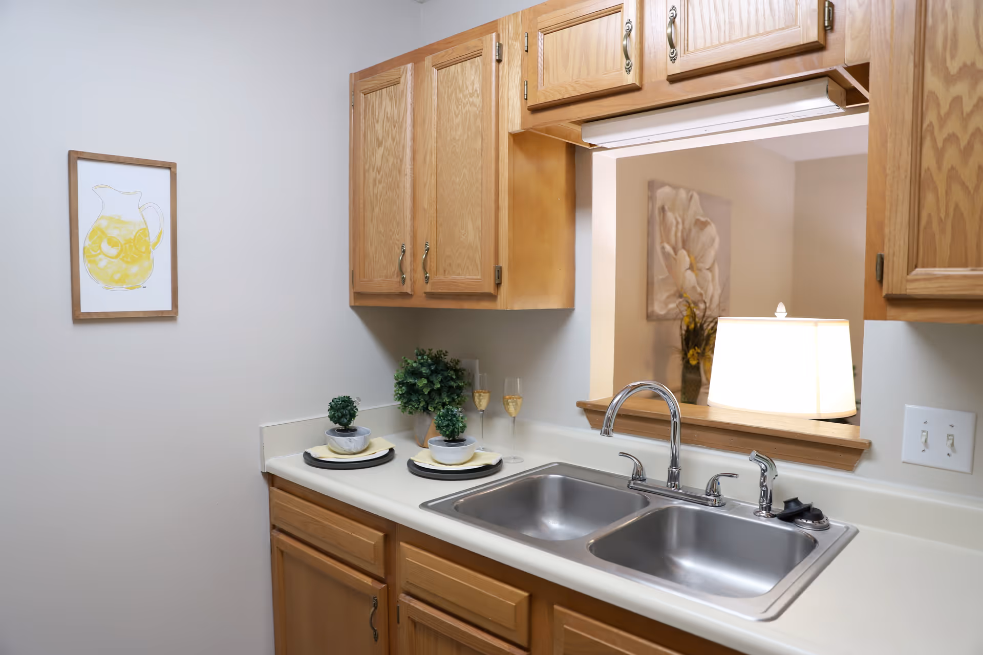 A kitchen area with wooden cabinets, a double stainless steel sink, and a countertop decorated with two small potted plants and two plates with bowls. There is a framed picture of a pitcher with lemon slices on the wall. A pass-through window reveals a lamp and a floral painting in the adjacent room.
