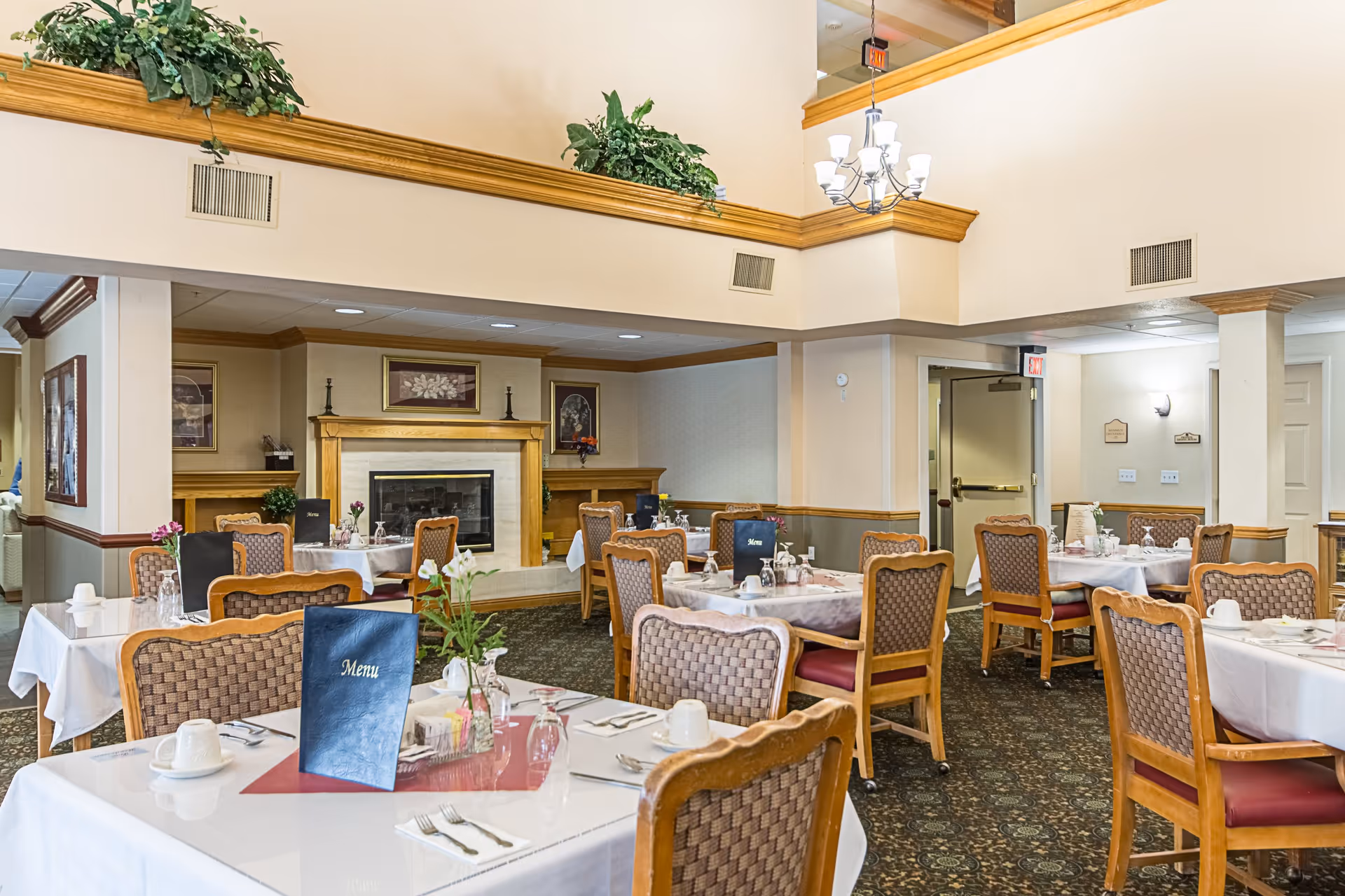 A dining room in a senior living facility with multiple tables covered in white tablecloths, each set with cups, utensils, and menus. The room features wooden chairs with woven backs, a carpeted floor, a fireplace with framed artwork above it, and plants on a ledge near the ceiling. There is a chandelier hanging from the ceiling and an exit door in the background.
