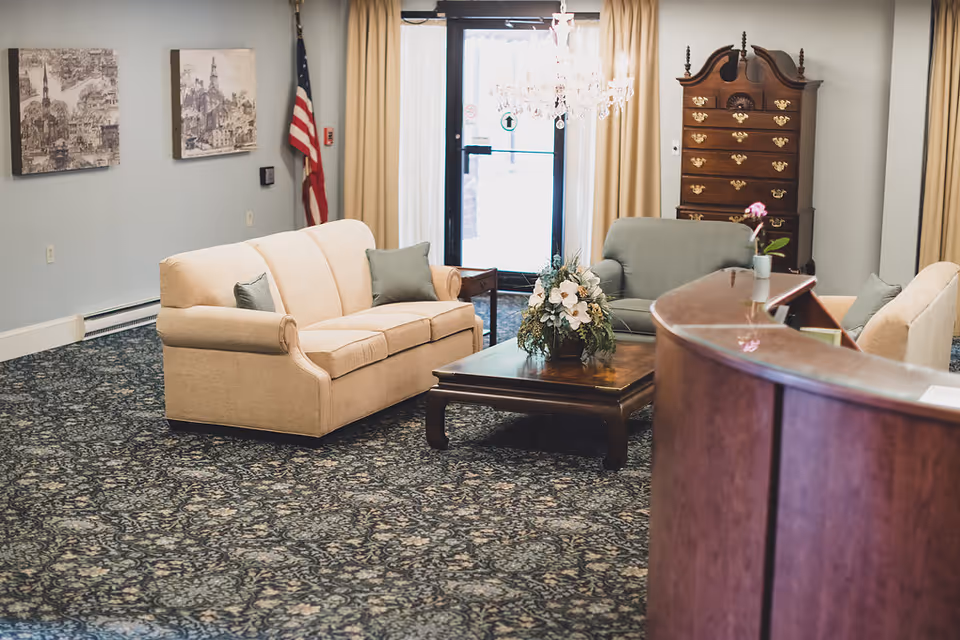 A cozy senior living center lounge area with beige and green sofas arranged around a wooden coffee table adorned with a floral centerpiece. The room features patterned carpet, a wooden reception desk in the foreground, an American flag, two framed black and white cityscape artworks on the wall, a chandelier, and a tall wooden chest of drawers near the entrance door with beige curtains.