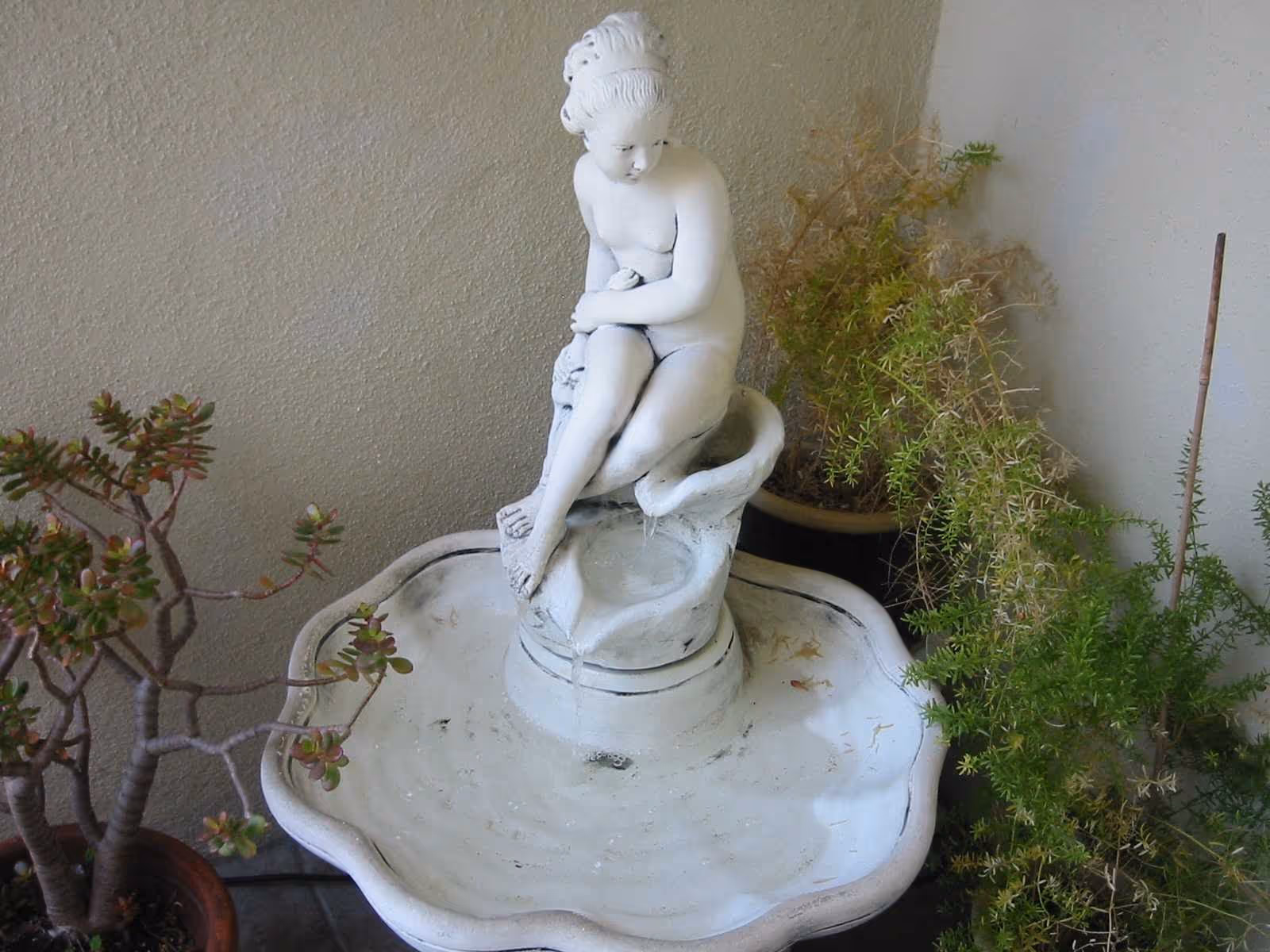 A white decorative water fountain featuring a statue of a seated woman with water flowing from her foot into a scalloped basin, surrounded by potted plants against a plain wall.