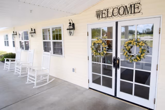 Covered porch area with white rocking chairs lined up along a cream-colored wall with windows. Double glass doors decorated with yellow floral wreaths and a black metal 'WELCOME' sign above the doors. Outdoor wall lanterns are mounted beside the windows and doors.