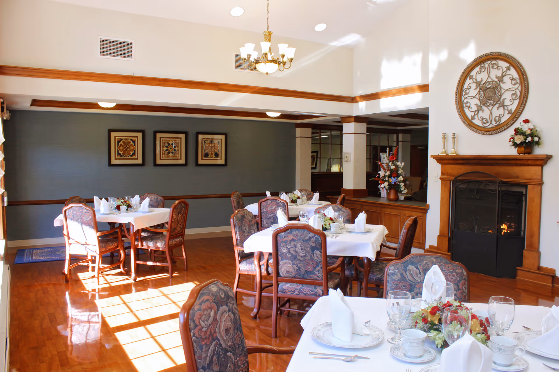 A bright dining room with wooden floors and several tables covered with white tablecloths, set with plates, glasses, and napkins. The chairs have floral upholstery. There is a fireplace with a decorative metal wall hanging above it and floral arrangements on a side table. The room has a chandelier and framed artwork on the walls.