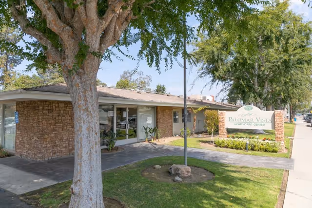 Exterior view of Palomar Vista Healthcare Center showing a single-story building with brick and beige walls, large windows, a sidewalk, green lawn, trees, and a sign with the facility name near the street.