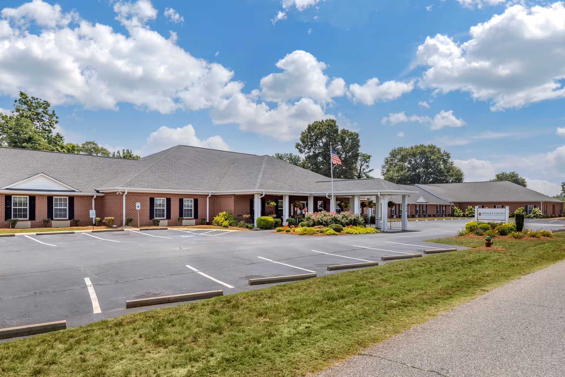 Exterior view of Wesley Court Assisted Living Community showing a single-story brick building with a gray roof, an American flag on a flagpole, landscaped flower beds, and an empty parking lot under a partly cloudy blue sky.