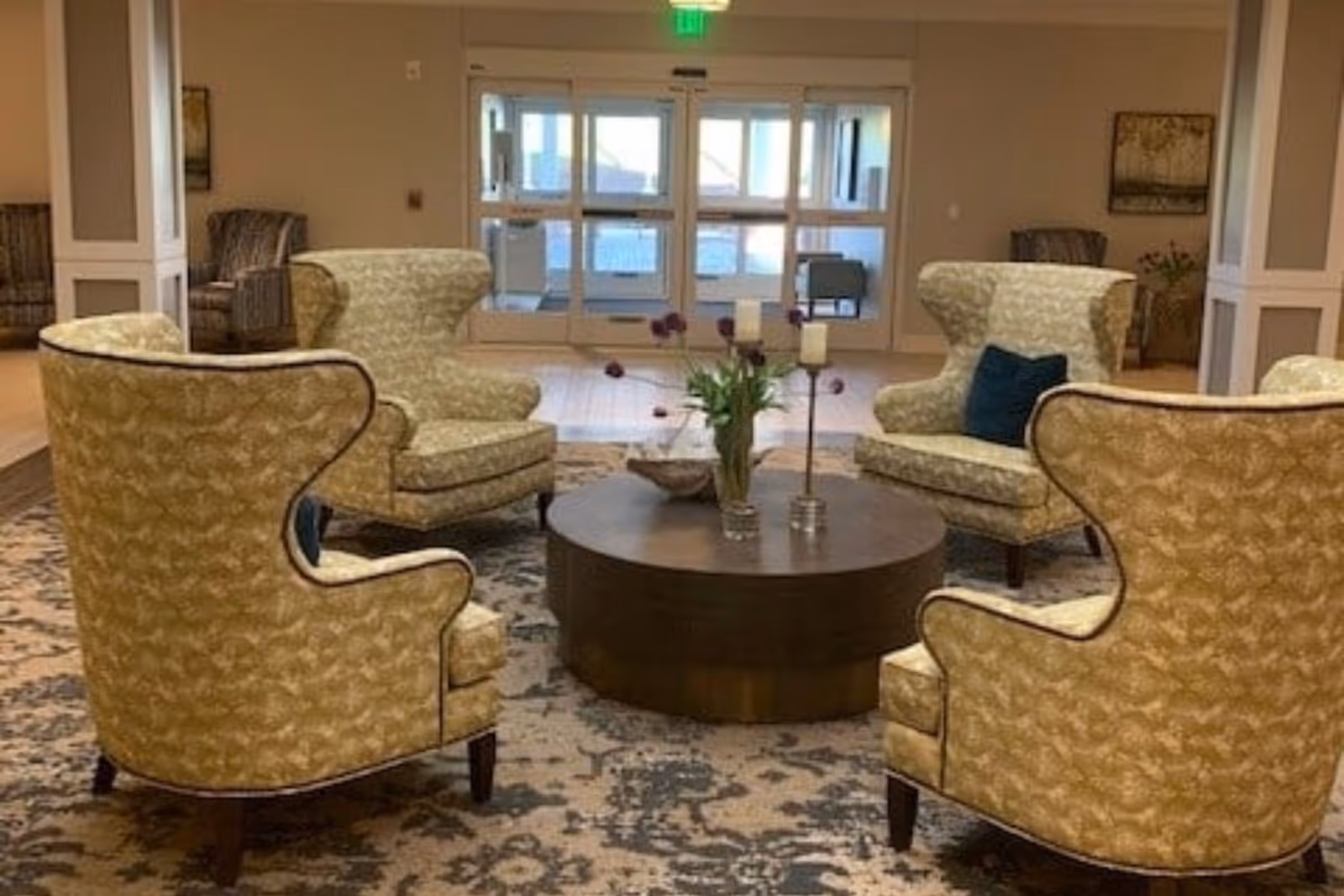 Lobby seating area with four patterned wingback chairs arranged around a round wooden coffee table in front of glass entrance doors.