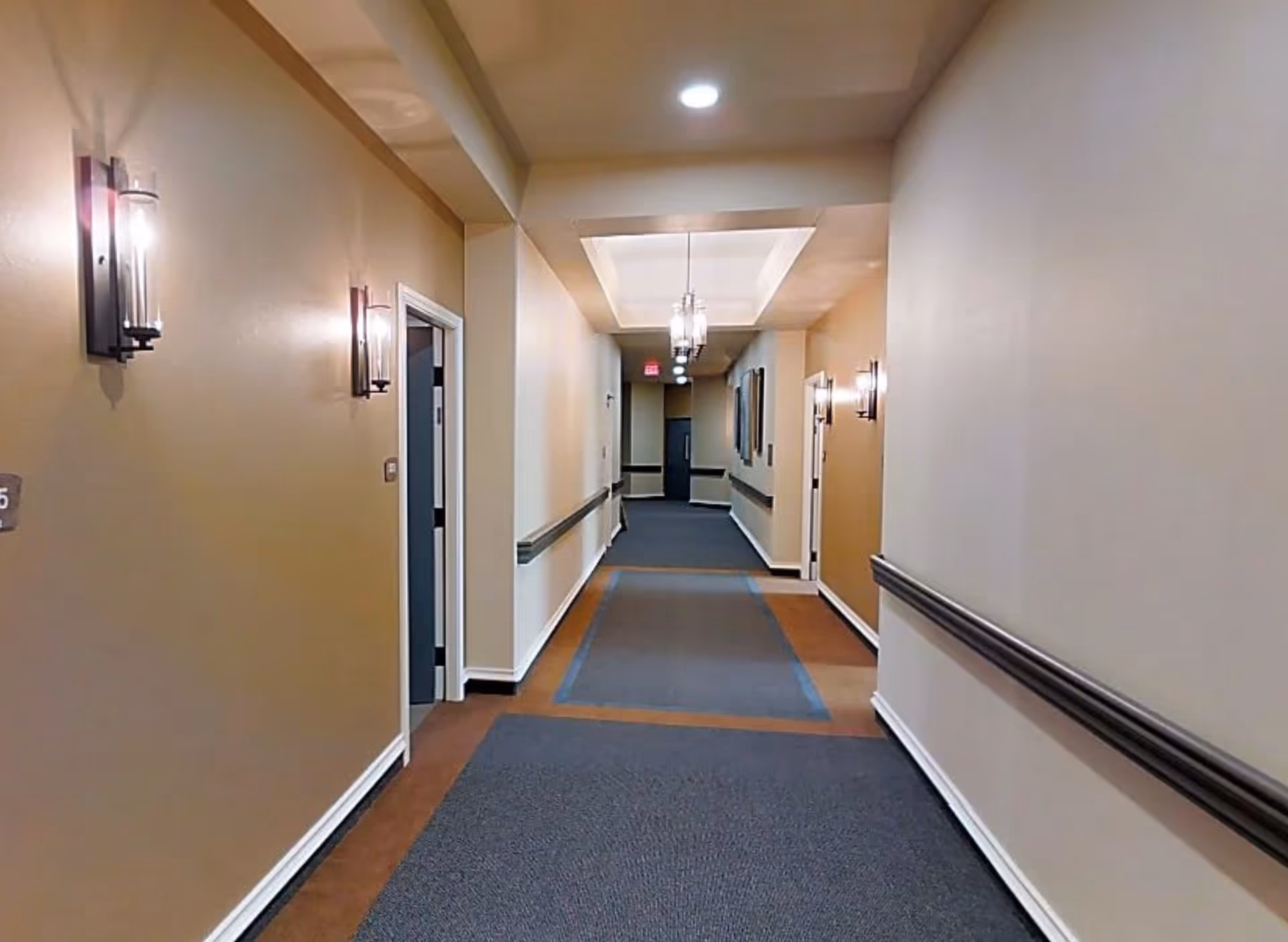 A well-lit indoor hallway with beige walls and dark carpeted flooring. The hallway has wall-mounted lights on both sides and handrails along the walls. Several doorways are visible along the corridor, and ceiling lights hang from a recessed ceiling section.