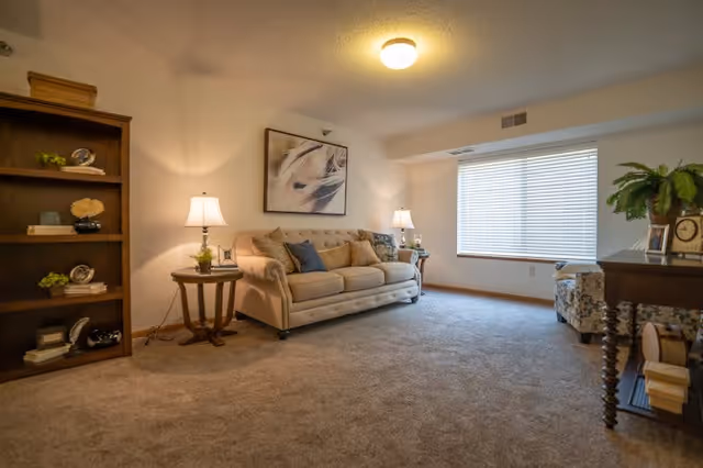 A cozy living room with a beige tufted sofa adorned with multiple cushions, two side tables each with a lamp, a wooden bookshelf with decorative items, a framed abstract artwork on the wall, a large window with blinds, a patterned armchair, and a wooden console table with a clock and a potted plant.