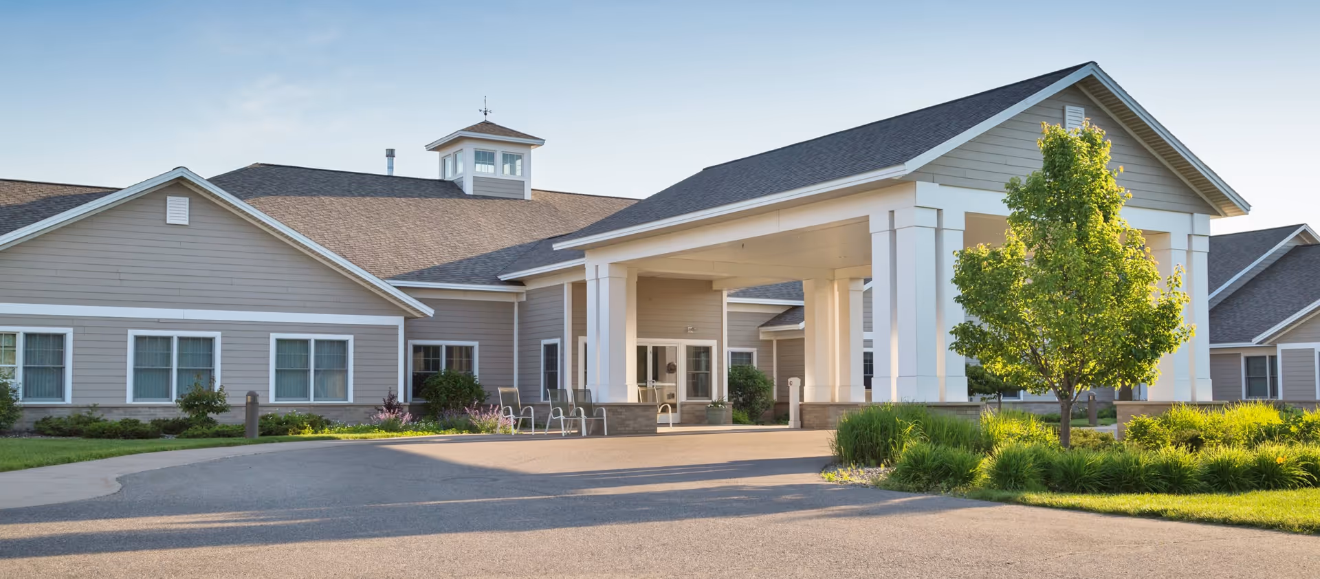 Exterior view of Mill Creek Senior Living Community building with a covered entrance, beige siding, multiple windows, and a landscaped area with green grass and a tree in front.