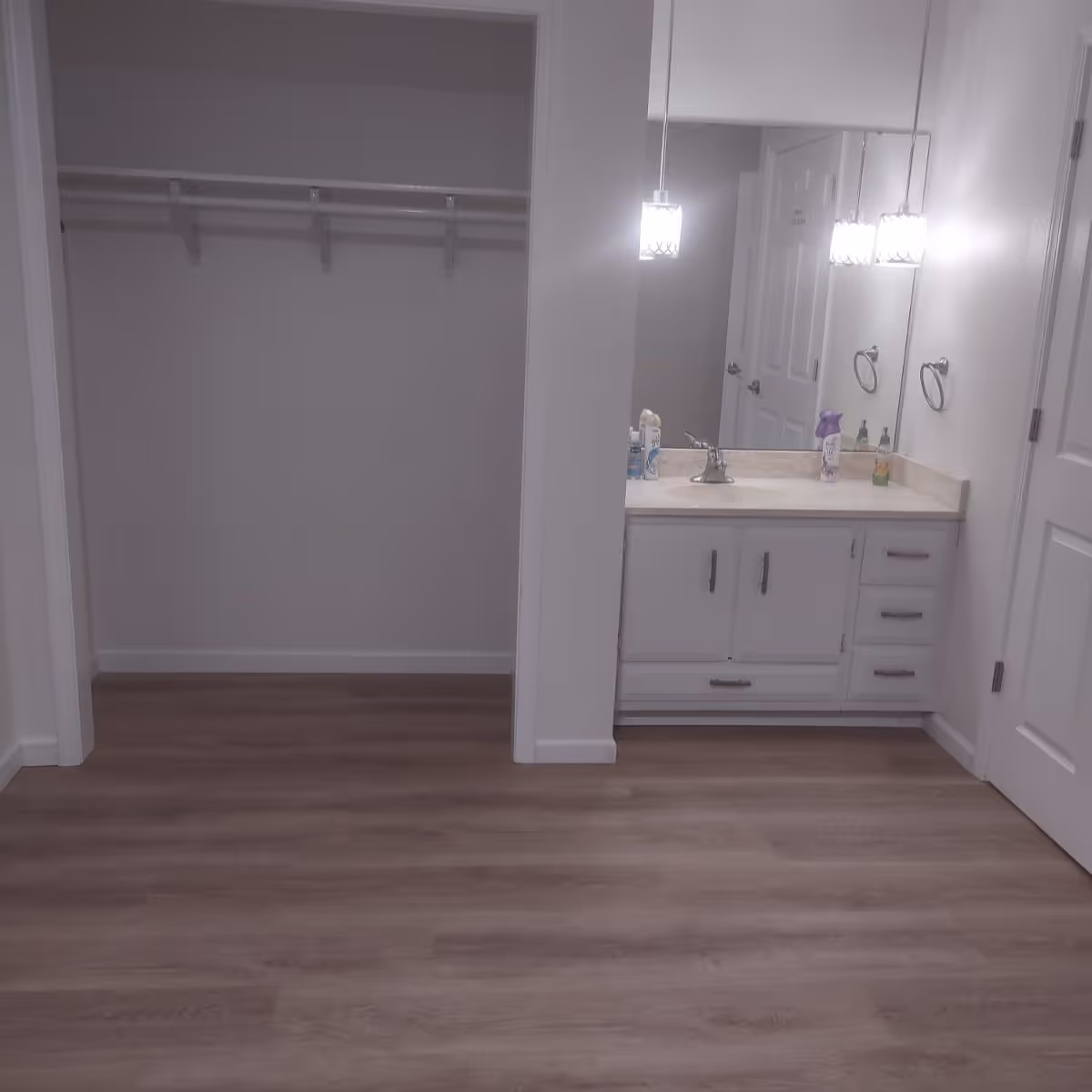 Interior view of a room with a wooden floor, an open closet with a hanging rod, and a bathroom vanity with a sink, mirror, and hanging light fixtures. Various toiletries are placed on the vanity countertop.